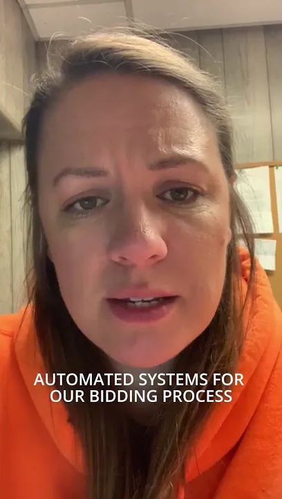 Video still: a woman in a high-visibility orange hooded top speaks to camera in a timber-panelled office. A caption burned into the video reads "automated systems for our bidding process".